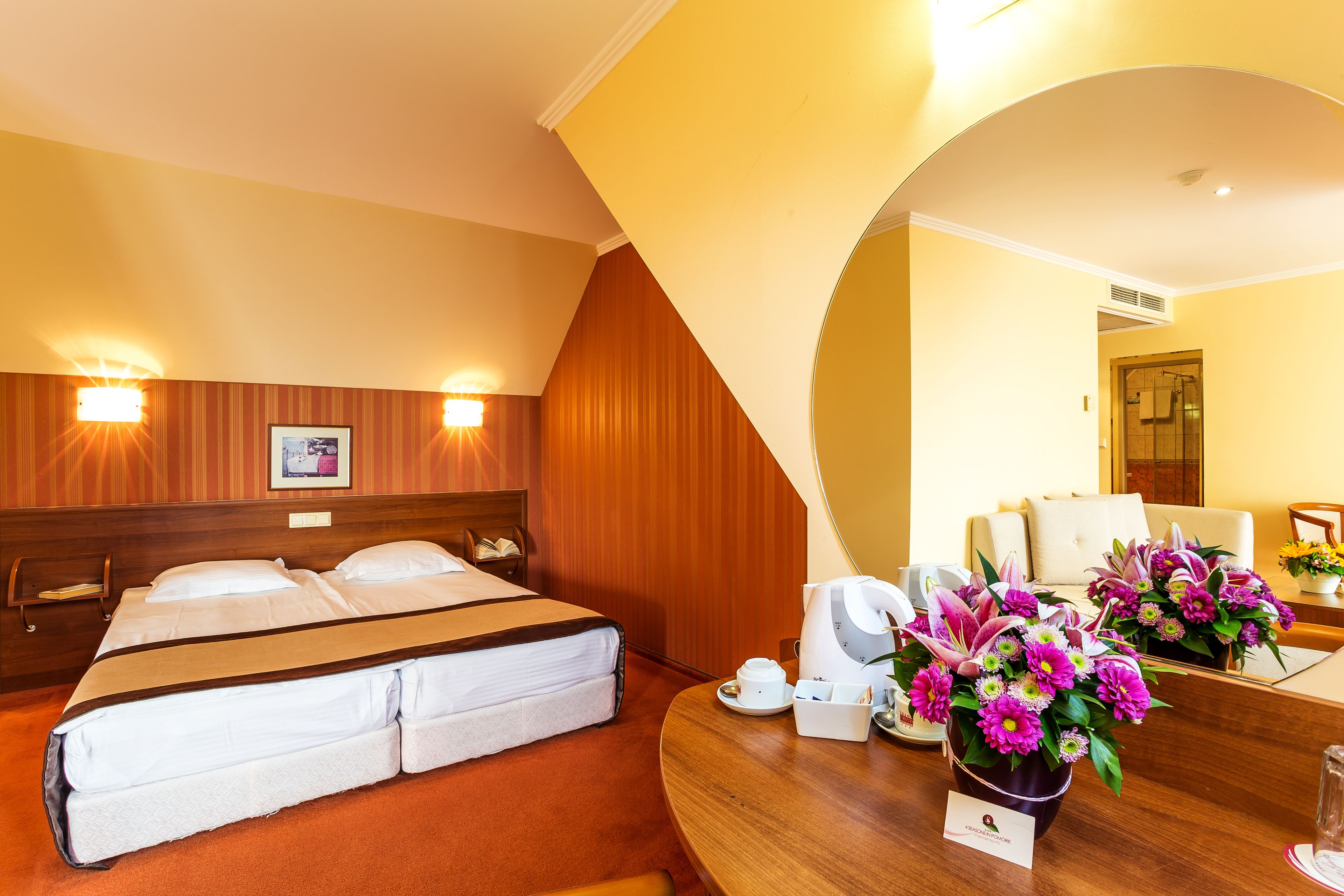 A hotel room with a double bed, bedside tables, a coffee maker, a vase of flowers, and a bathroom visible through an archway.
