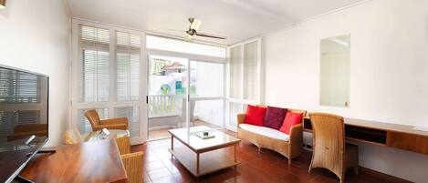 A spacious living area with a wooden desk, a sofa with red cushions, and a large window with blinds.