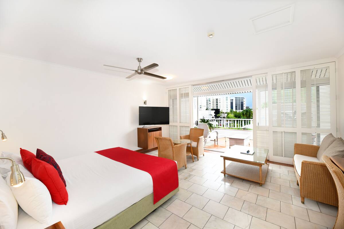 A hotel room with a bed, a TV, a balcony with outdoor furniture, and a view of buildings.