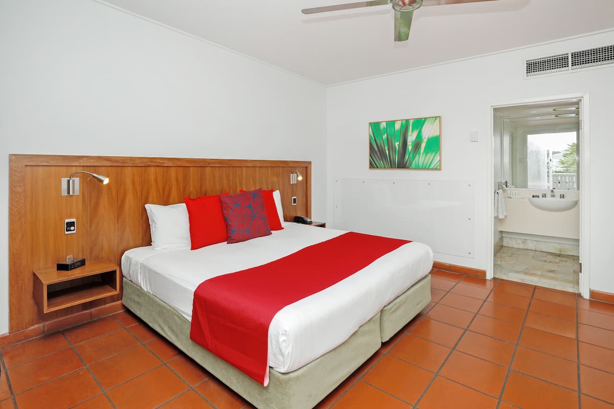 A hotel room with a wooden headboard, a large bed with white and red bedding, a red throw blanket, a nightstand with a lamp, a painting on the wall, and a bathroom with a sink and mirror.