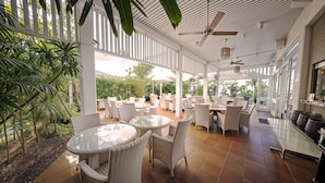 Ein Terrassenbereich im Freien mit runden Tischen und Korbstühlen, umgeben von weißen Geländern und viel Grün.