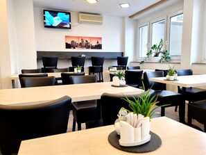 A modern dining area with multiple tables and chairs, a flat-screen TV mounted on the wall, and a cityscape painting above the tables. - Adler Hotel Frankfurt (Frankfurt)