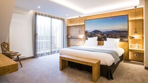 A modern hotel room with a large bed, a wooden headboard, a painting of a mountain landscape, a bench, and a desk with a lamp. - Hostellerie des Châteaux & Spa (Ottrott)