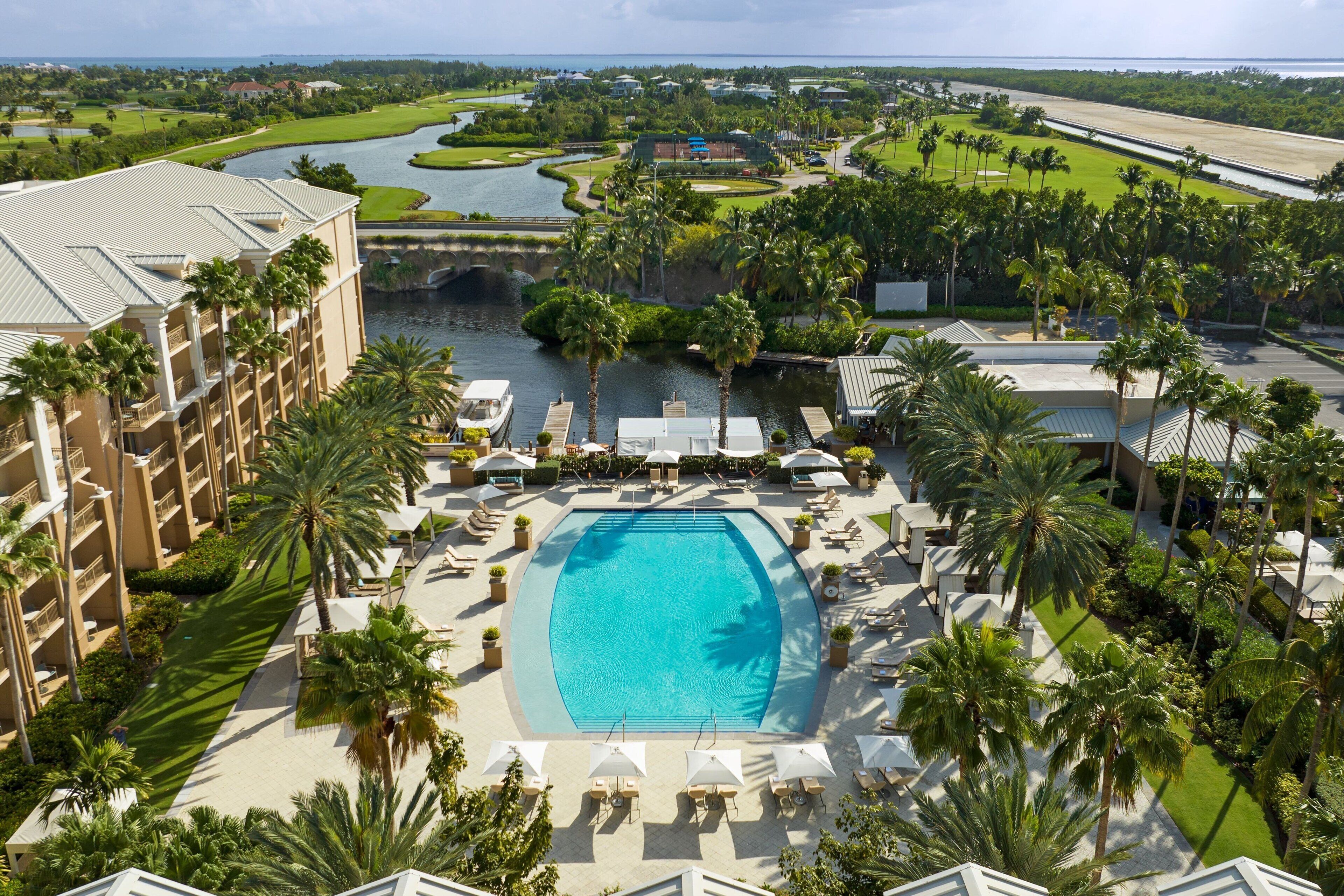 Un resort con piscina central, rodeado de palmeras y sillas de descanso, y un campo de golf y un canal de agua al fondo.