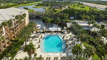 Un resort con piscina central, rodeado de palmeras y sillas de descanso, y un campo de golf y un canal de agua al fondo.