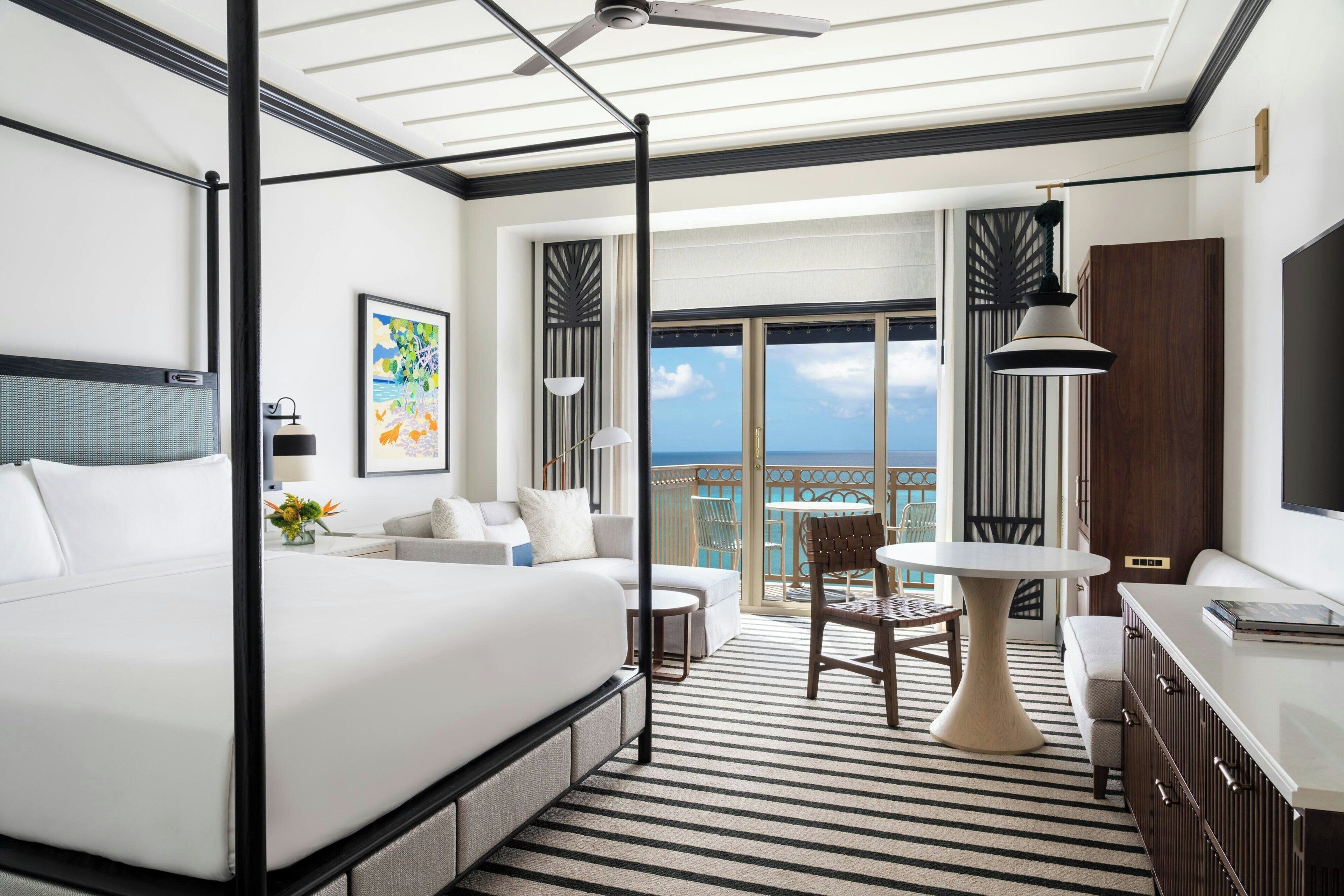 Una habitación de hotel moderna con una cama grande, un balcón con vistas, una zona de comedor y un televisor.