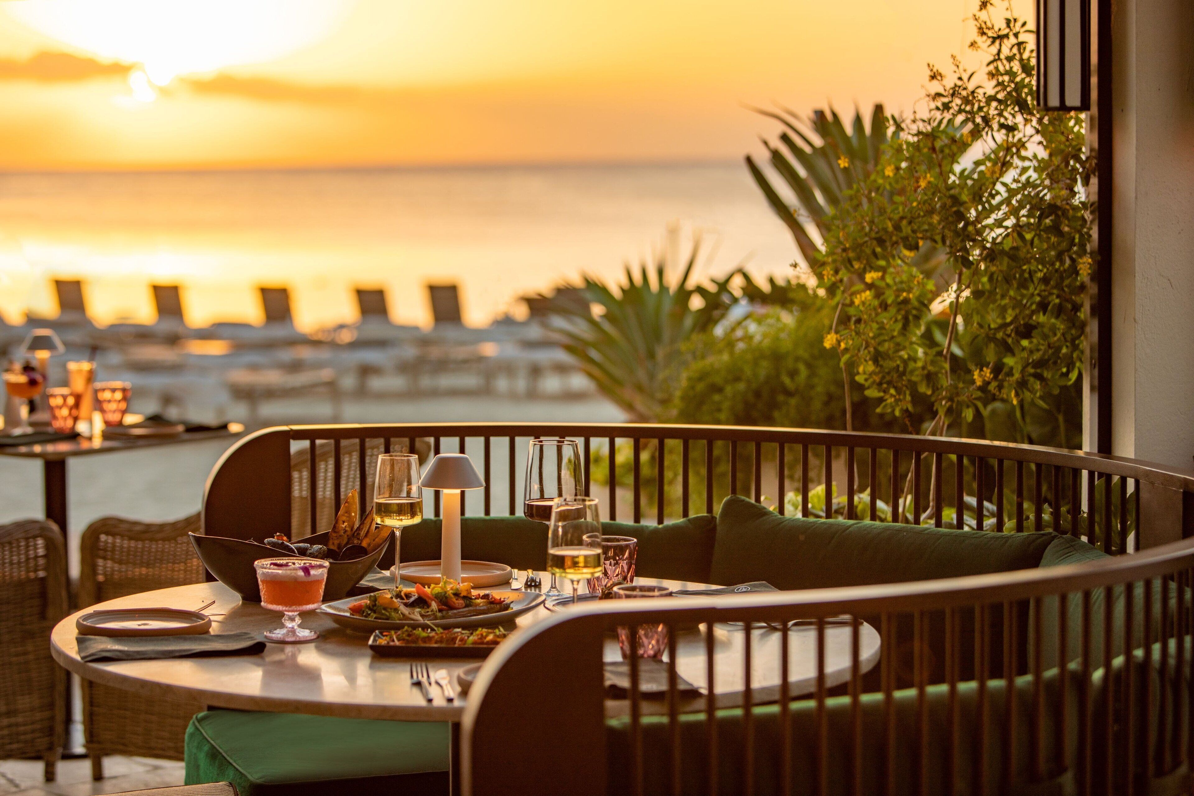 A dining table with a view of the sunset over the ocean, surrounded by chairs and a bench.