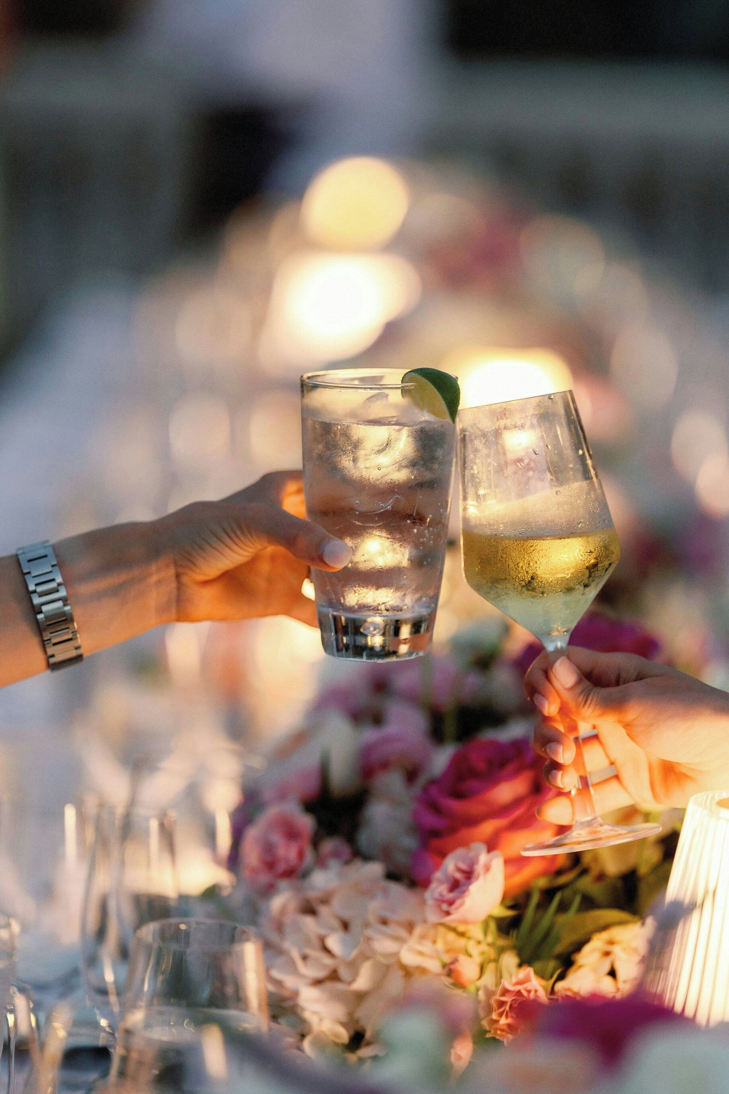 To hænder holder glas med drikkevarer, med en sløret baggrund af et bordopdækning og blomster.