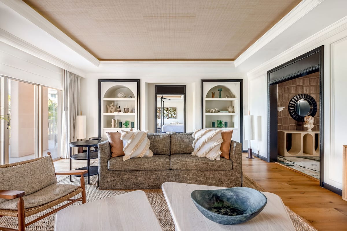 A spacious living room with a large sofa, a coffee table, and a fireplace. There are shelves with decorative items and a large mirror on the wall.
