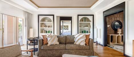 Un salón amplio con un sofá grande, una mesa de centro y una chimenea. Hay estantes con objetos decorativos y un espejo grande en la pared.