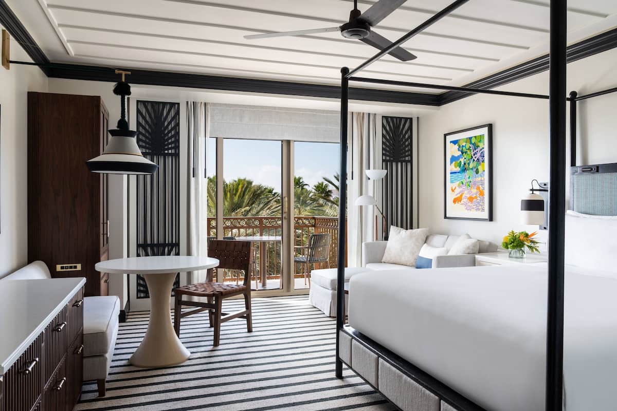 A hotel room with a large bed, a desk, a chair, a table, and a balcony with a view of palm trees.