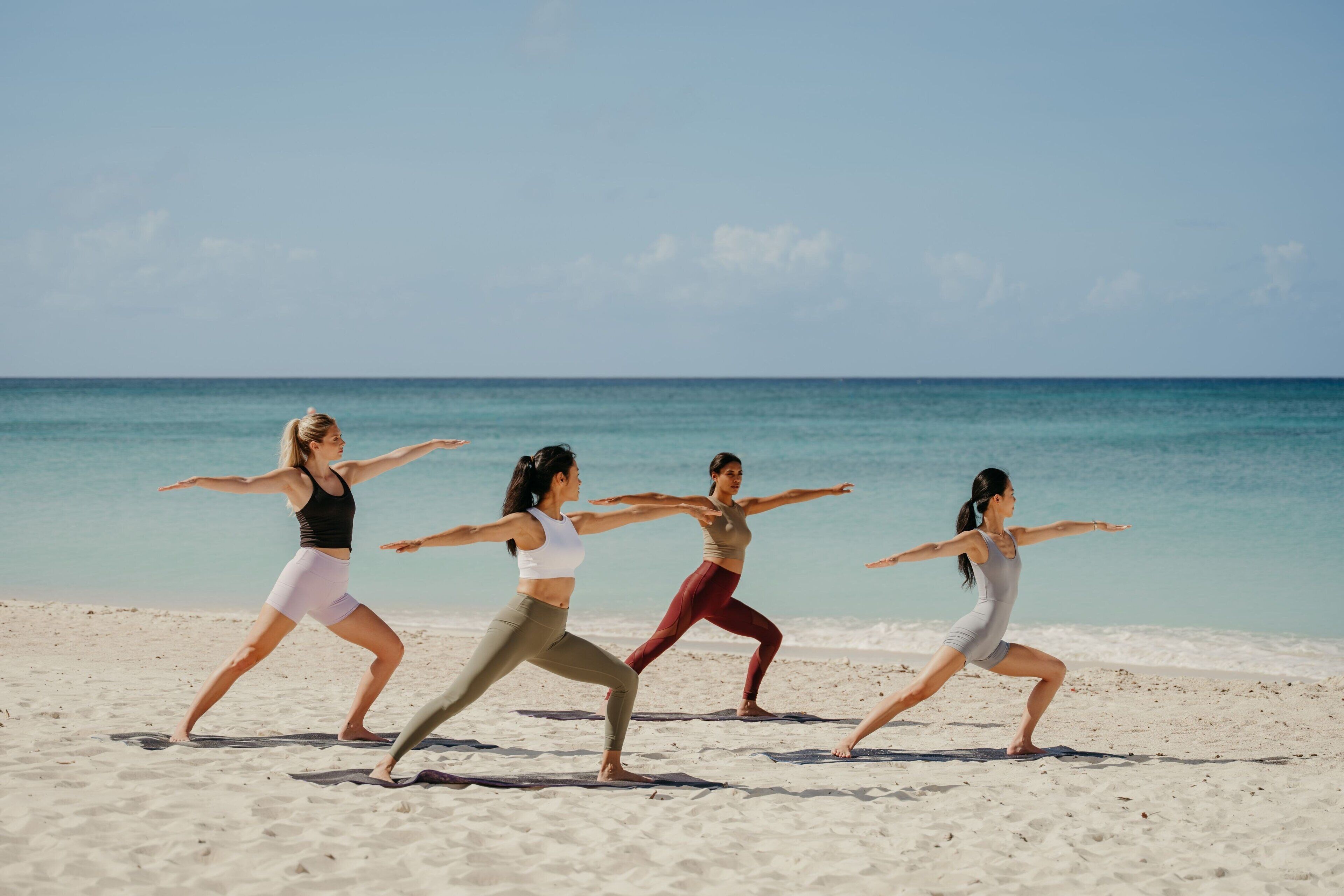 Four individuals performing a synchronized yoga routine on a sandy beach with a clear sky and calm sea in the background.