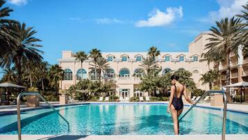 A swimming pool with a person standing at the edge, surrounded by palm trees and a building with arched windows in the background.