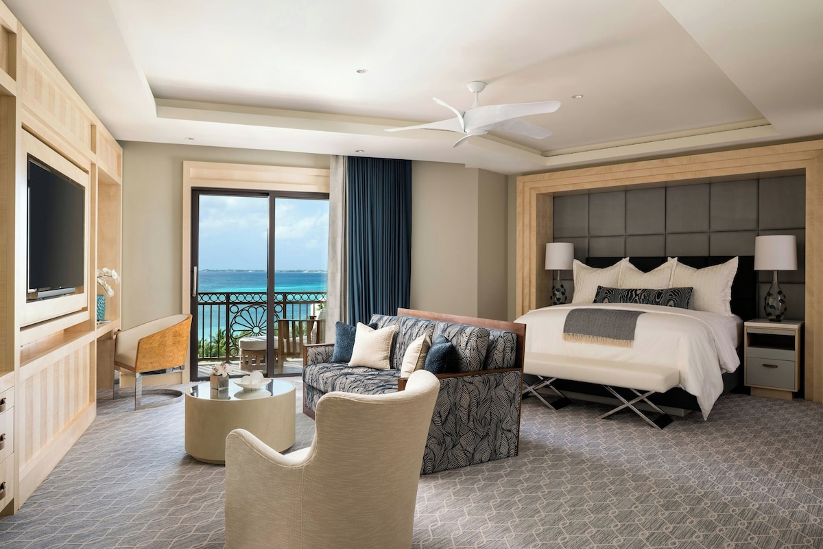 A hotel room with a bed, a sofa, a TV, a balcony with a view of the sea, and a ceiling fan.