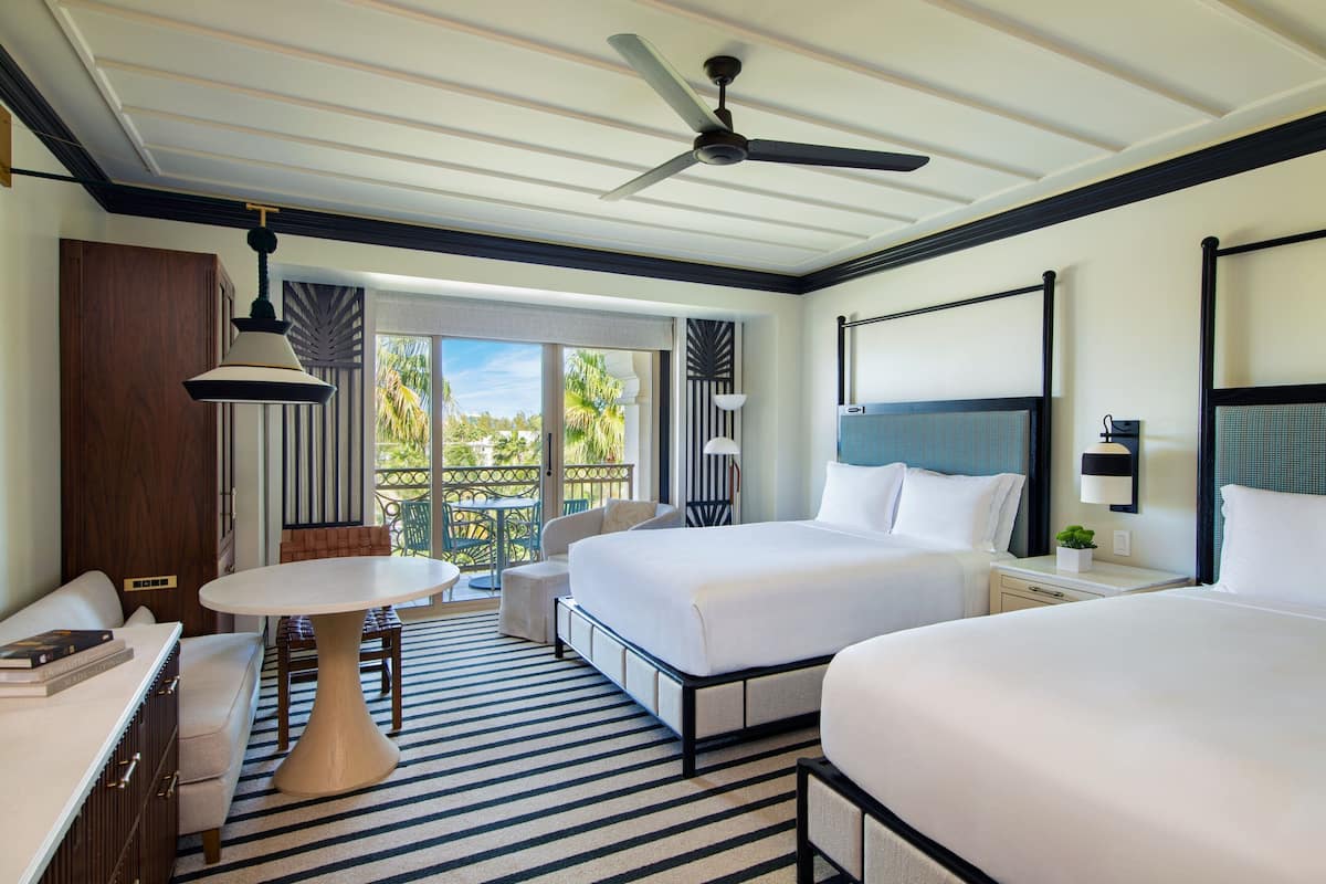 A hotel room with two beds, a dining table, a sofa, a ceiling fan, and a view of the outdoors.