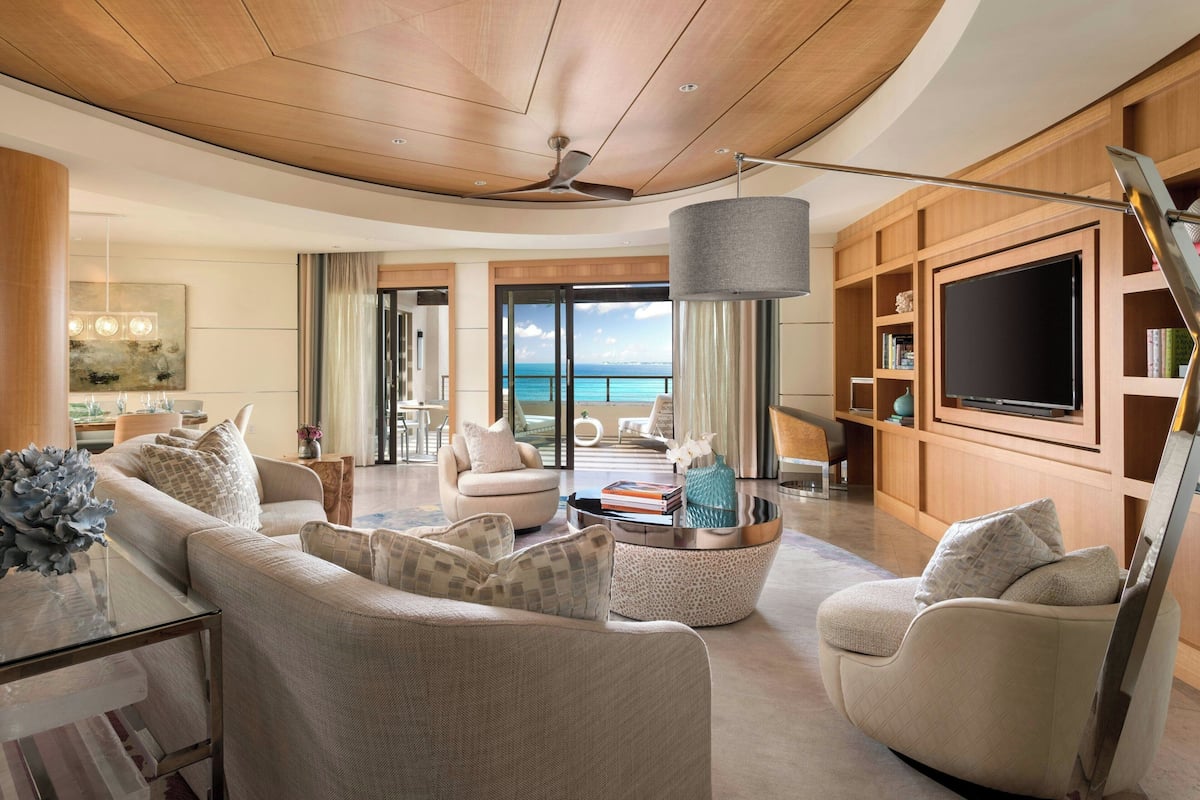 A spacious living room with a large TV, comfortable seating, a ceiling fan, and a view of the ocean.