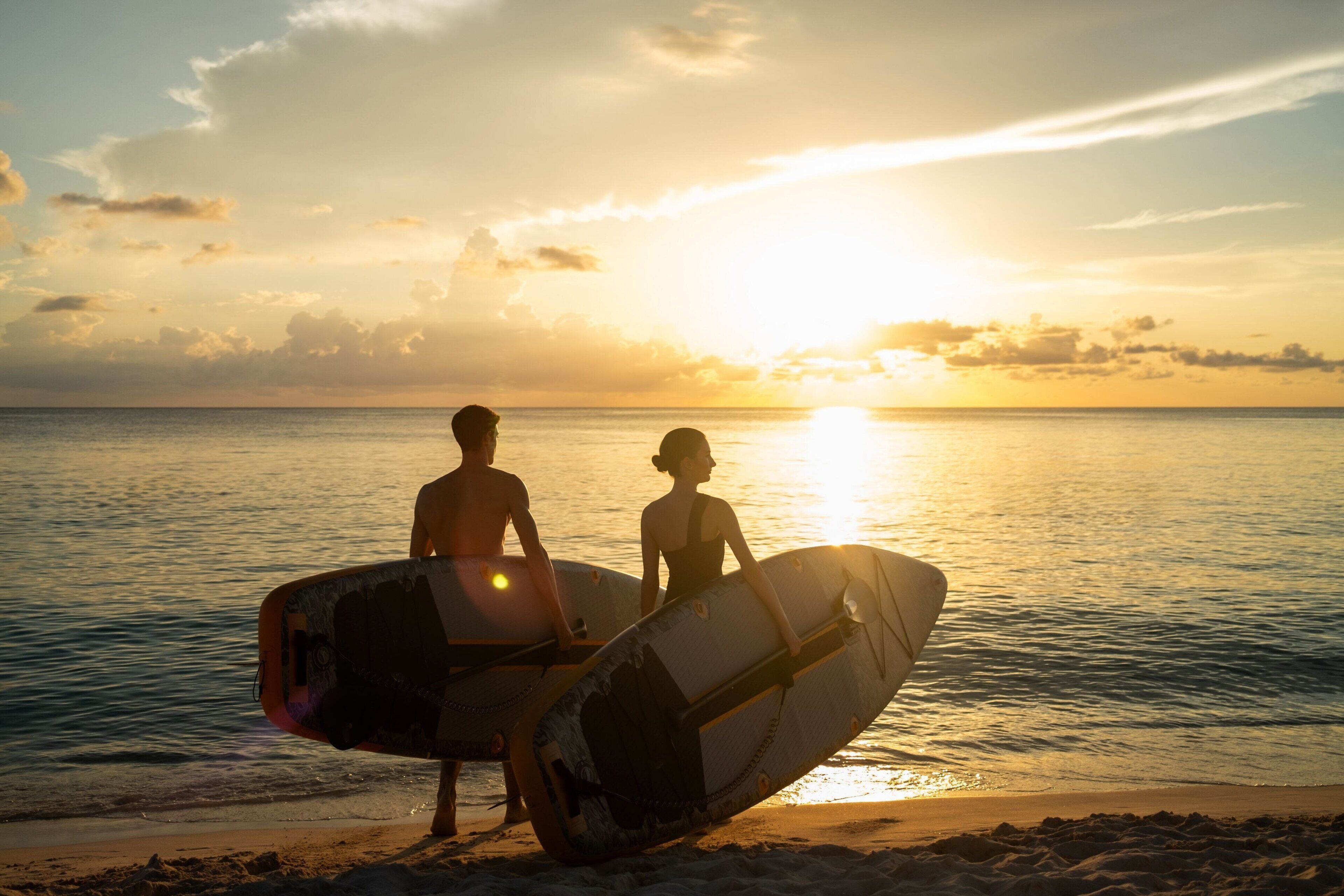 Two people carrying surfboards on a beach at sunset.