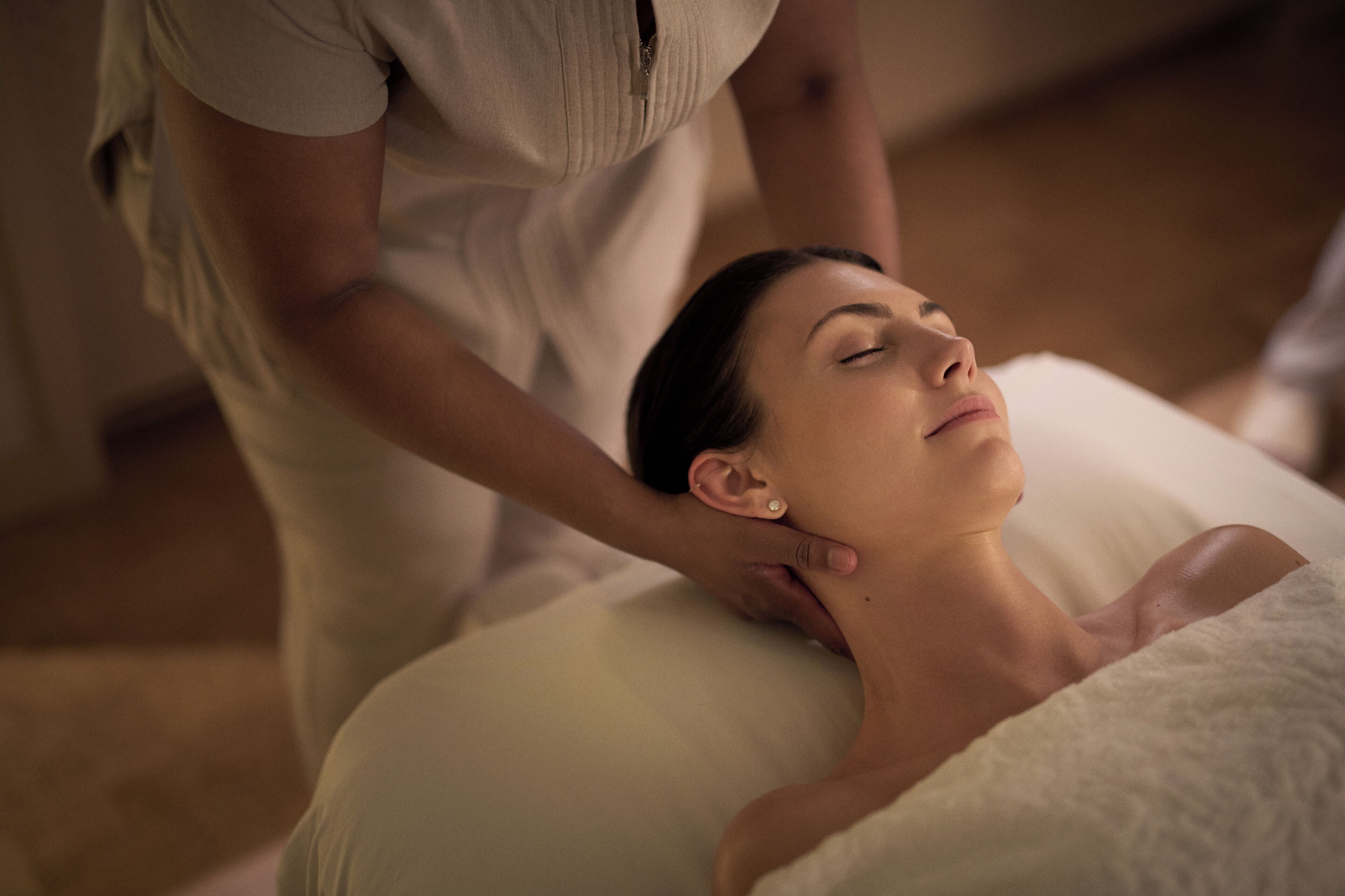 A person receiving a neck massage in a room with a white bed and a wooden floor.