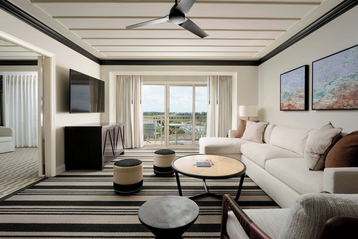 A modern hotel room with a flat-screen TV, a ceiling fan, a sofa, a coffee table, ottomans, and a view of the outdoors.