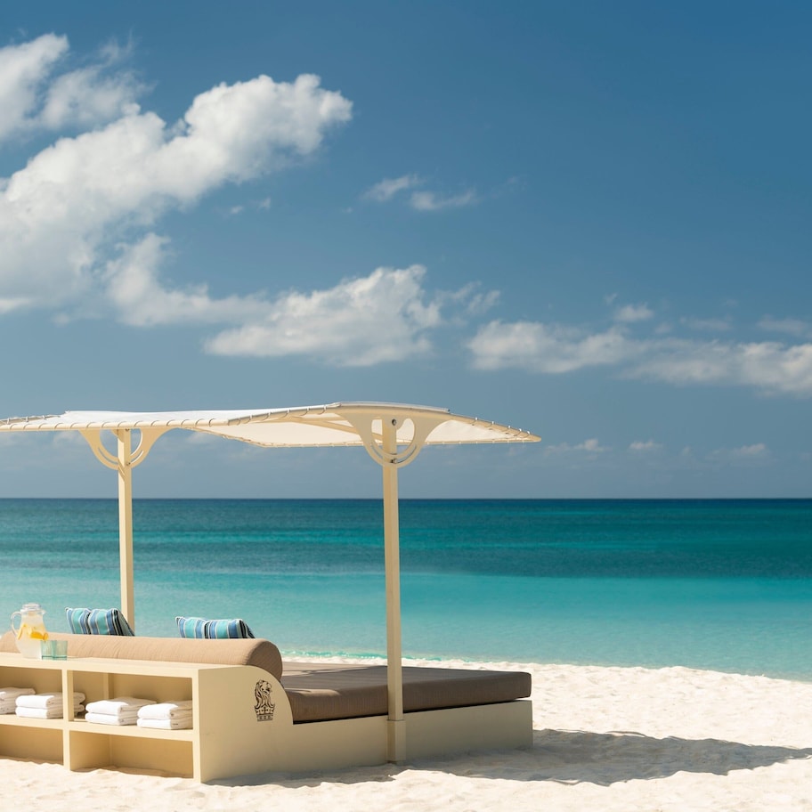 A beachside lounge with a cushioned seating area, a small table with a pitcher, and a canopy umbrella.