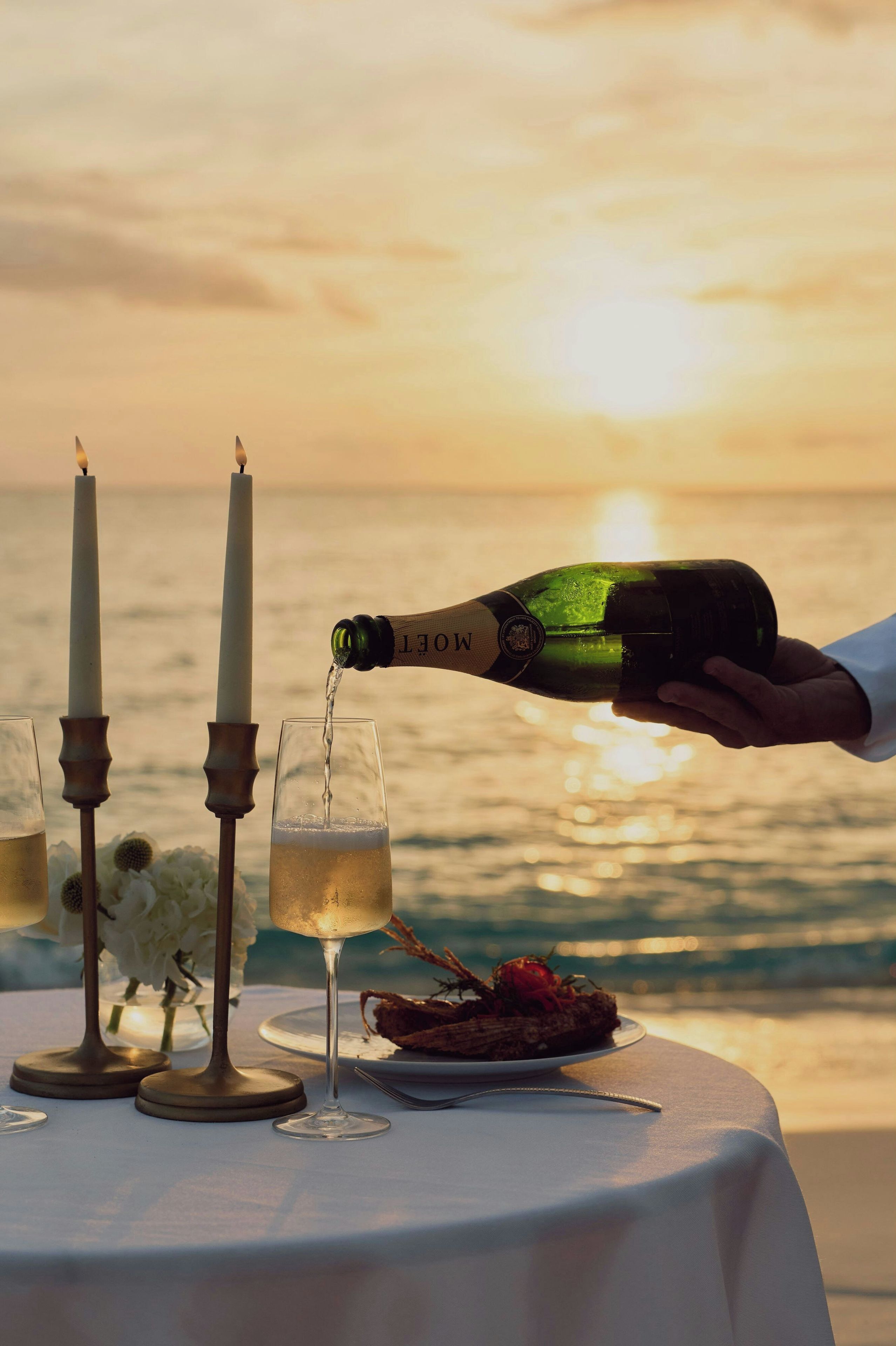 Botella de champán siendo servida en una copa, con una vela y flores sobre una mesa frente al mar.