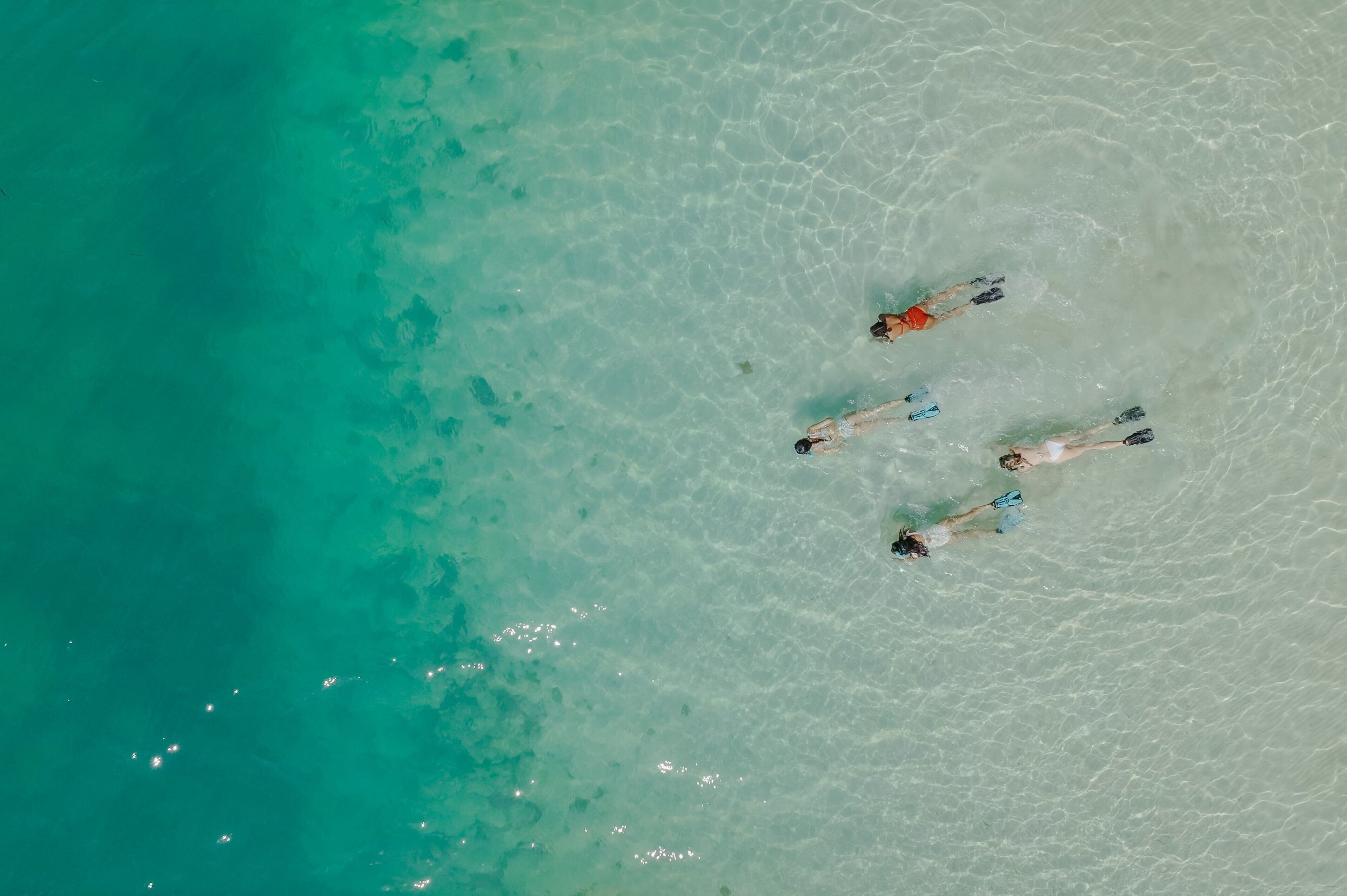 An aerial view of a beach with clear turquoise water and three people swimming.