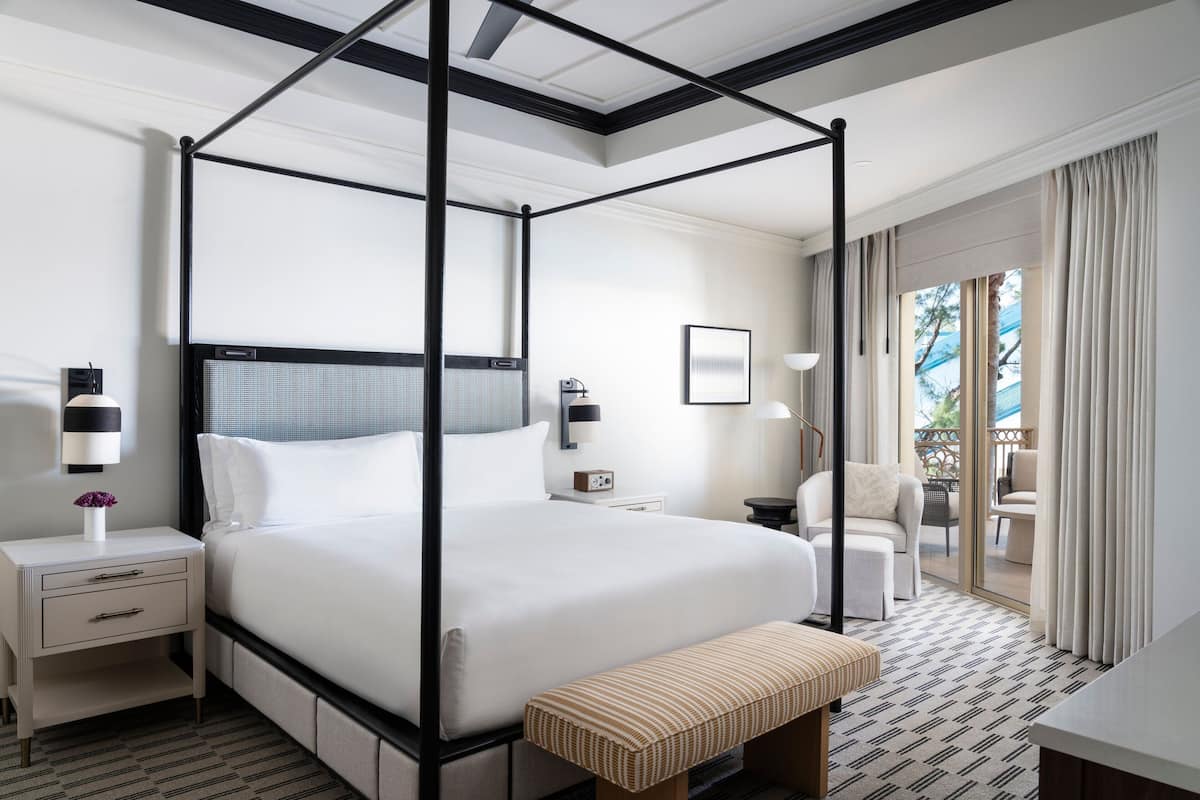 A four-poster bed with a canopy, a bedside table with a lamp, a chair, and a view of the outdoors through a sliding glass door.