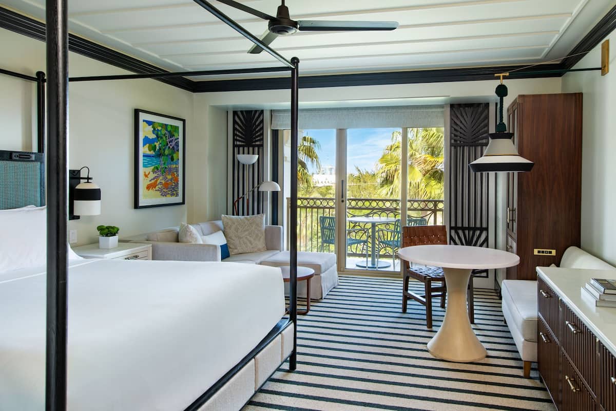A hotel room with a large bed, a sofa, a dining table, and a balcony with a view of palm trees.