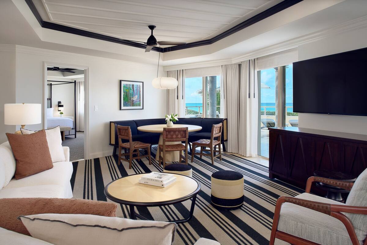 A hotel room with a balcony, a dining area, and a living room with a sofa, chairs, and a coffee table.