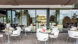A modern outdoor cafe with white chairs and tables, large windows, and a cobblestone floor. - Sotel Nomentana (Rome)