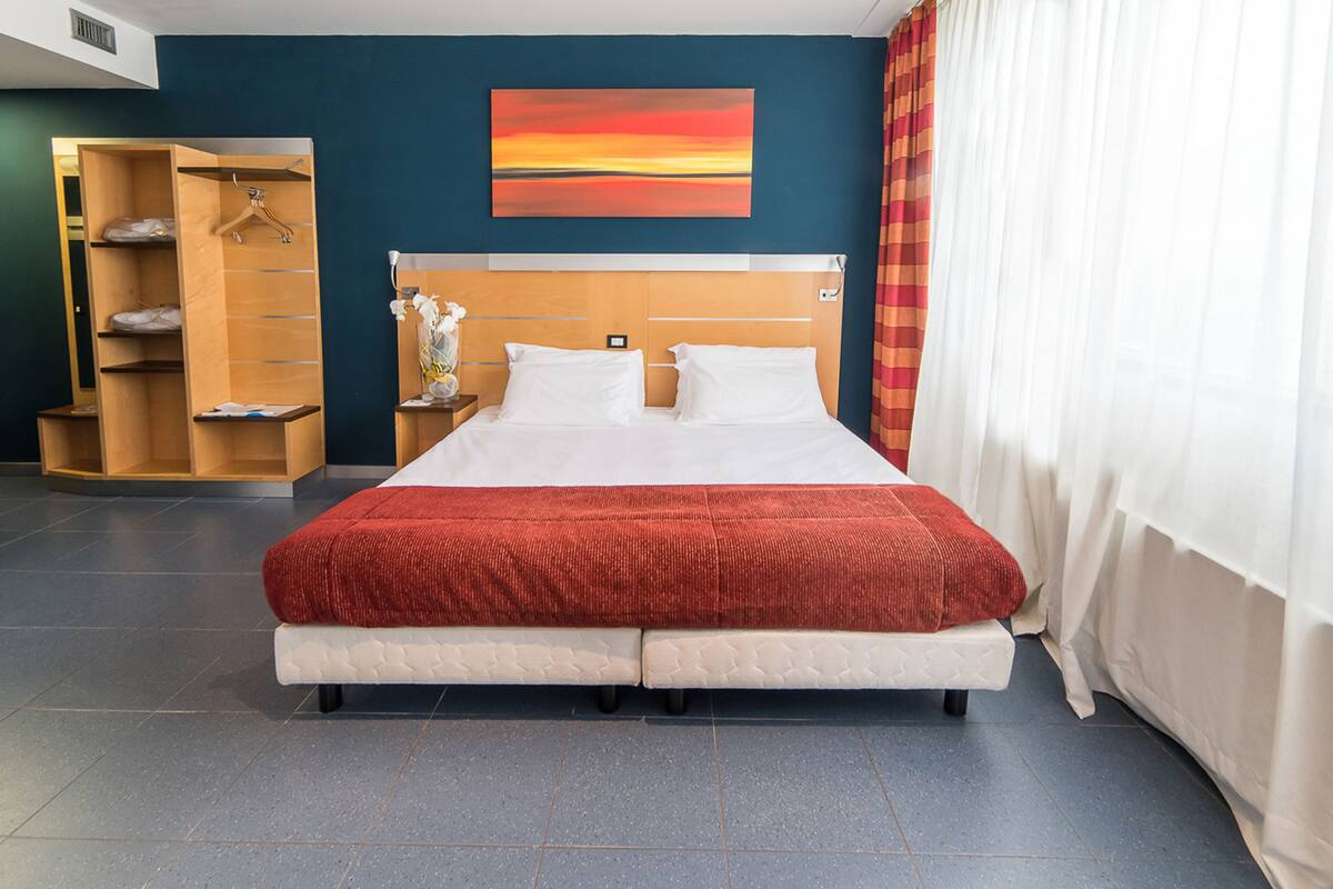 Habitación de hotel con una cama grande, un armario de madera, una mesita de noche con un florero y un cuadro en la pared.