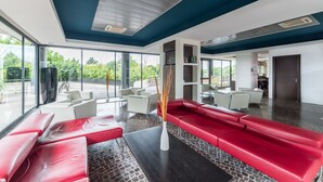A modern lobby with red seating, white tables, and large windows offering a view of greenery. - Sotel Nomentana (Rome)