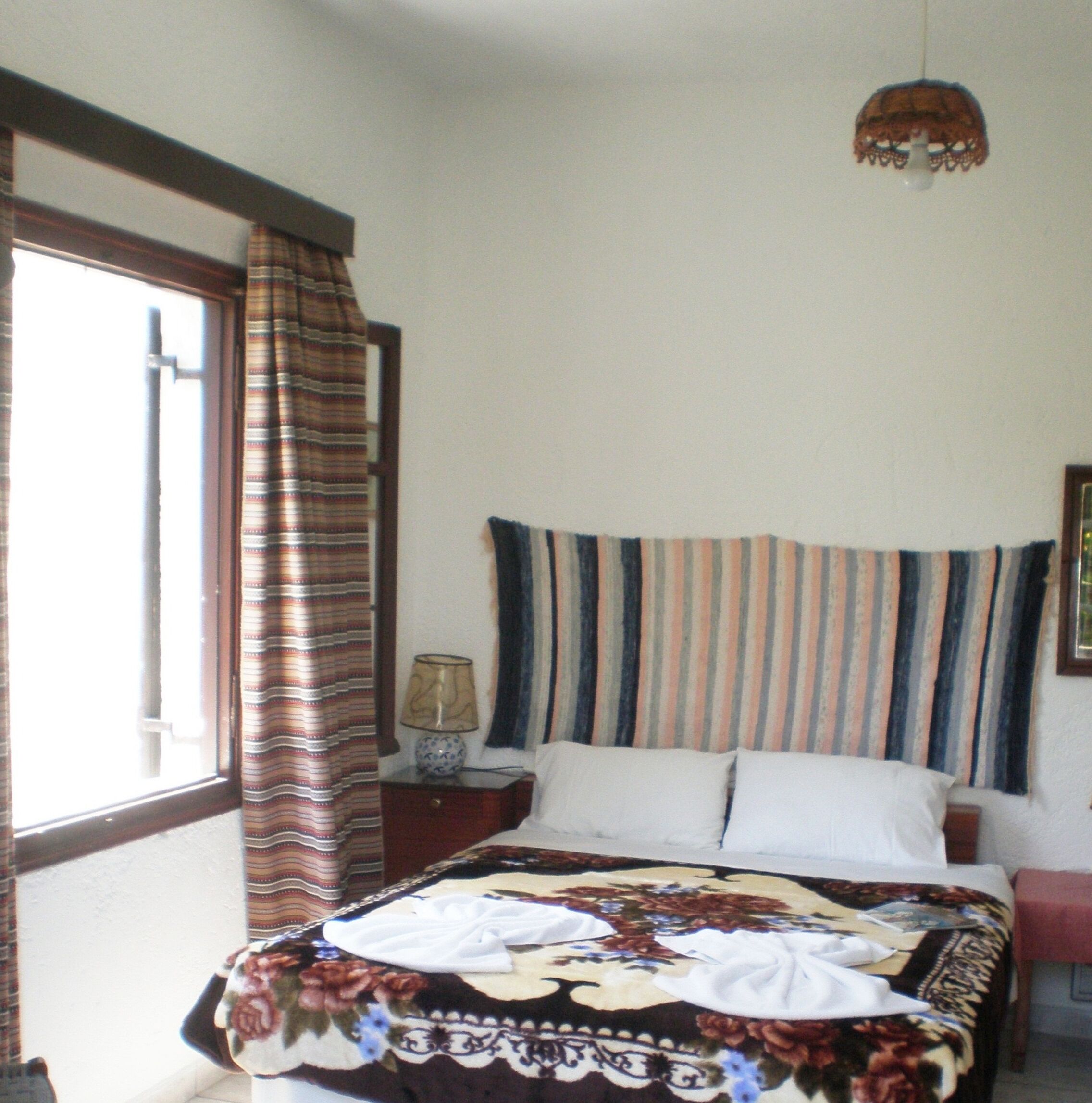 A bedroom with a bed, a nightstand, a lamp, a window with curtains, and a hanging light fixture.