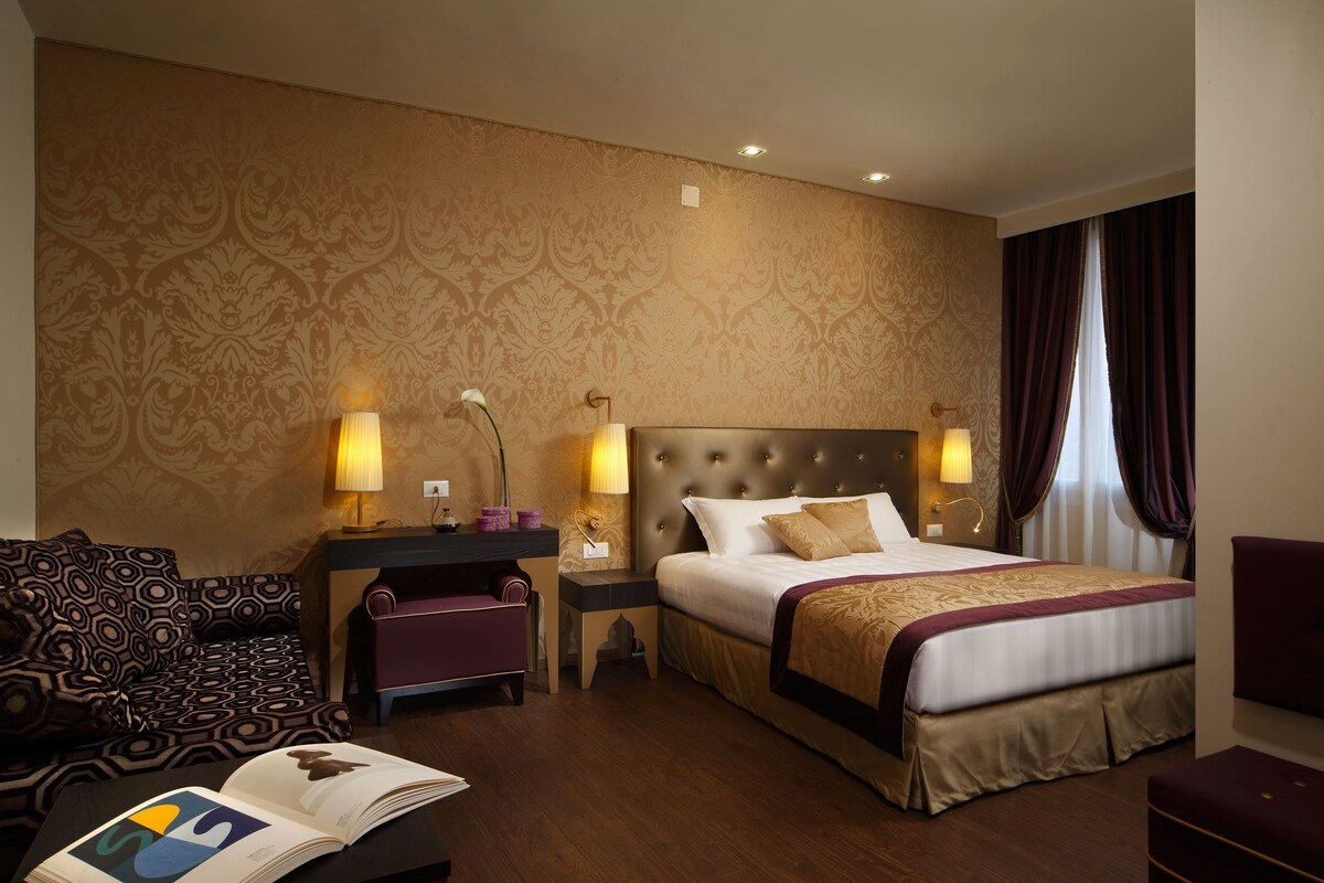 A hotel room with a large bed, two bedside tables with lamps, a sofa, and a small table with a book.