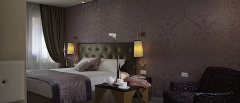 Habitación de hotel con cama, mesitas de noche, una silla y una pequeña mesa con una flor y una tetera.