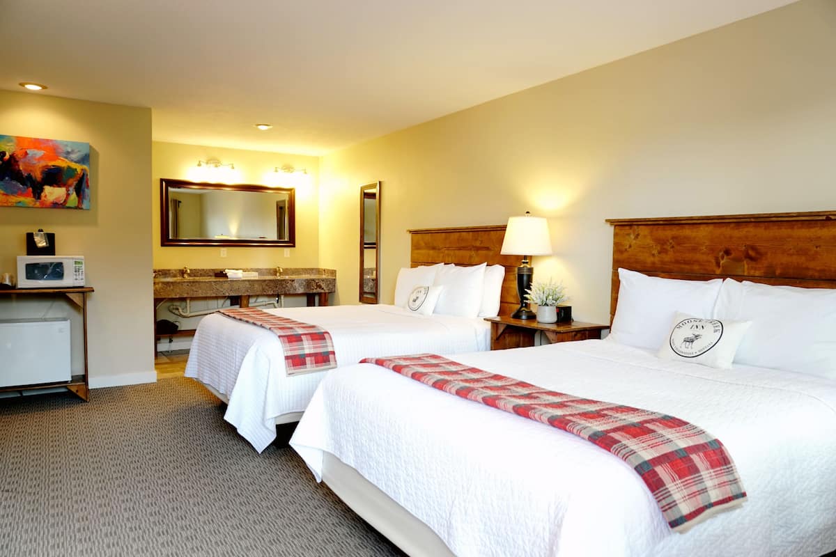 A hotel room with two beds, a large mirror, a wooden headboard, a bedside table with a lamp, and a small potted plant.