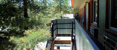 A wooden staircase leading to a balcony with a metal railing, surrounded by trees and a building with windows and a door.