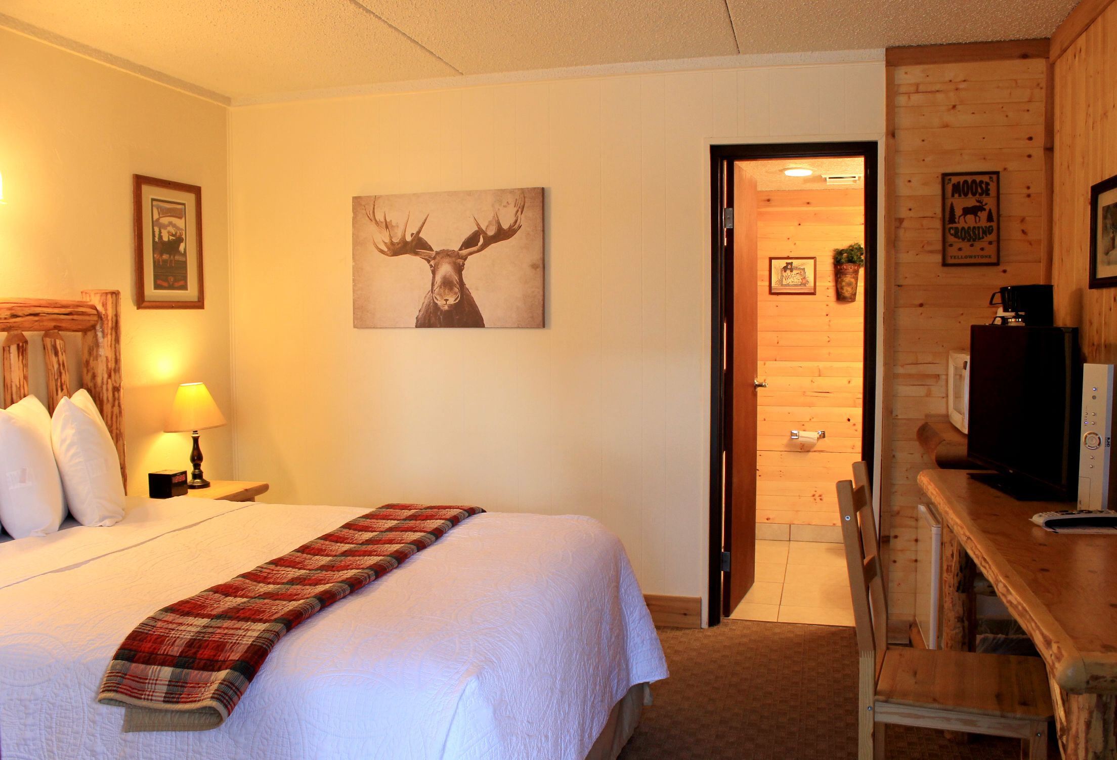 Una habitación de hotel con una cama, una manta escocesa, una obra de arte con un alce, un televisor y una pared de madera.