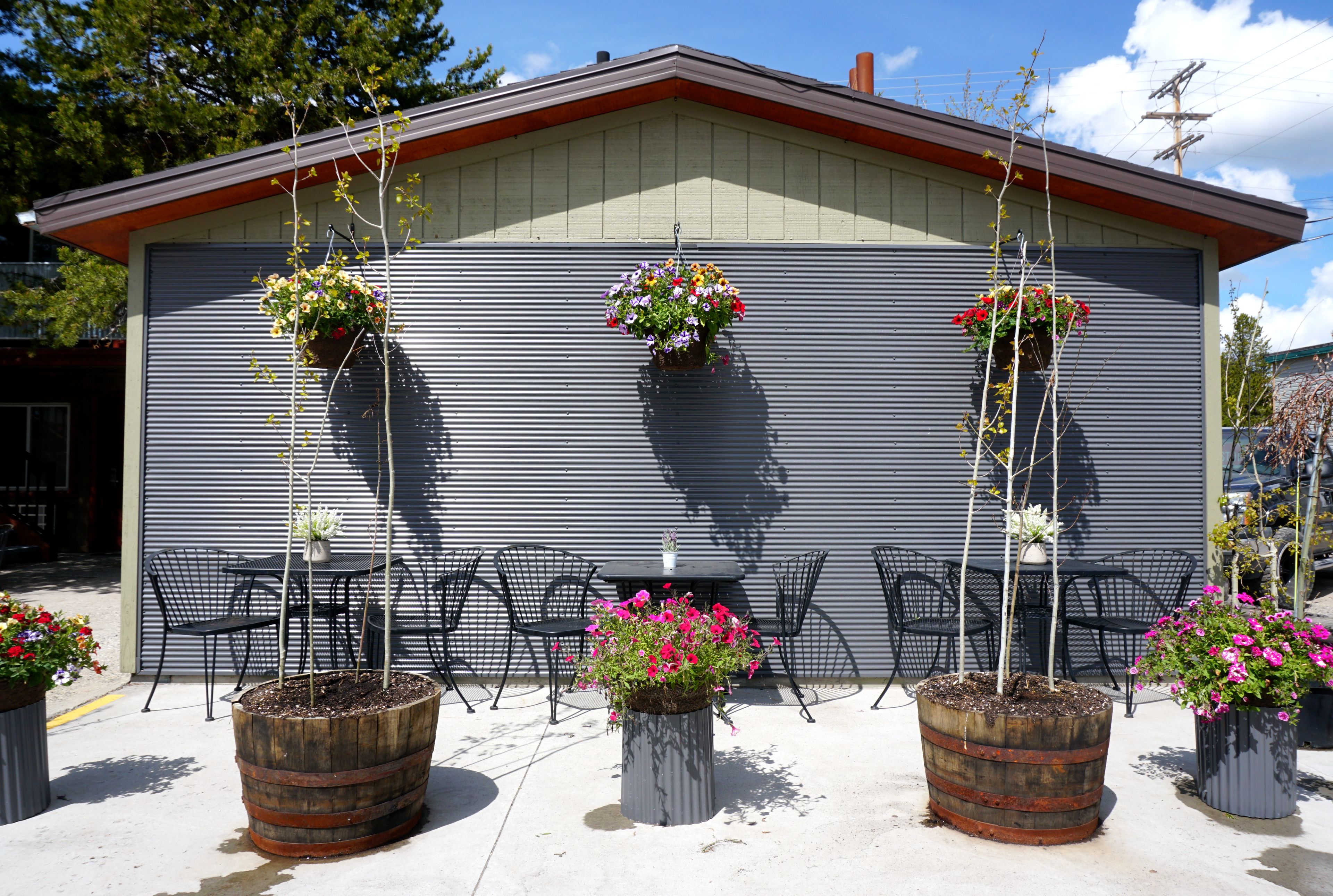 Un espacio al aire libre con sillas y mesas de metal, plantas en macetas y un edificio de fachada de metal corrugado.