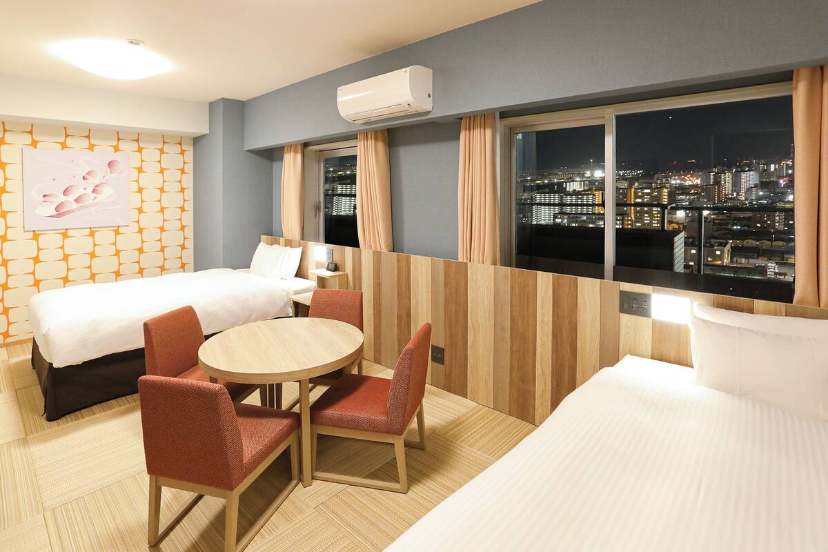 A hotel room with two beds, a dining table with chairs, a city view, and an air conditioning unit.