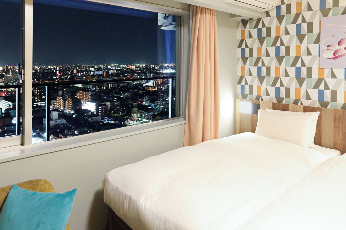 A hotel room with a large window offering a city view at night.