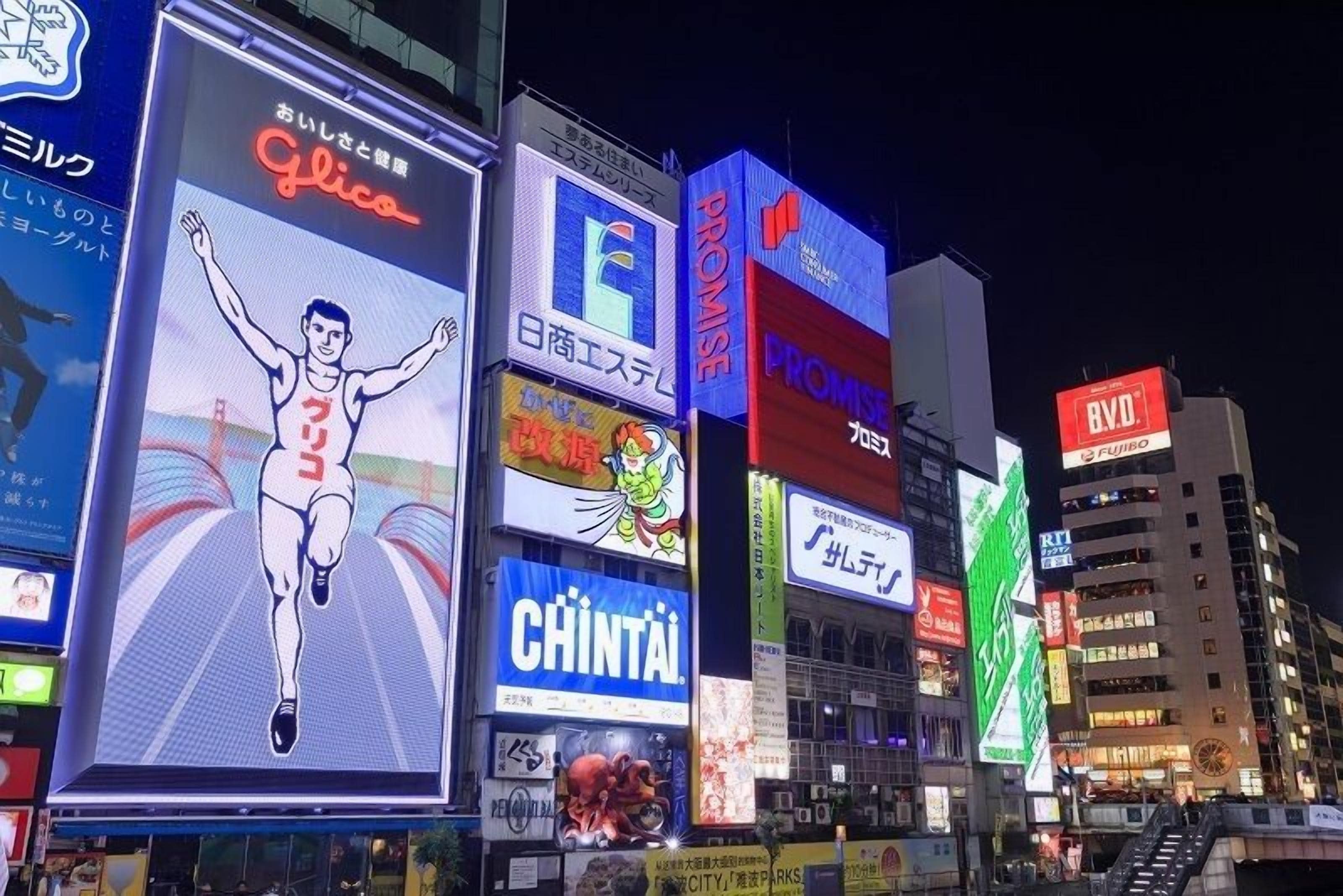 Vista nocturna de una zona urbana concurrida, con vallas publicitarias iluminadas y carteles que anuncian diversos negocios.