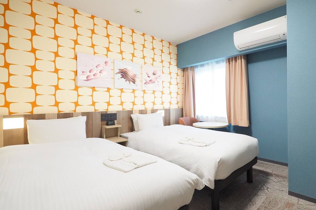 Une chambre d’hôtel avec deux lits, une table de chevet, un bureau, un climatiseur et une fenêtre avec des rideaux.