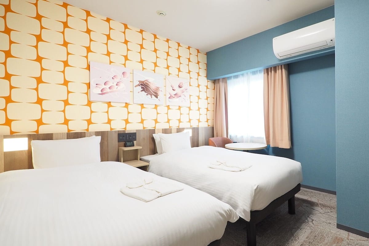 A hotel room with two beds, a nightstand, a desk, an air conditioner, and a window with curtains.