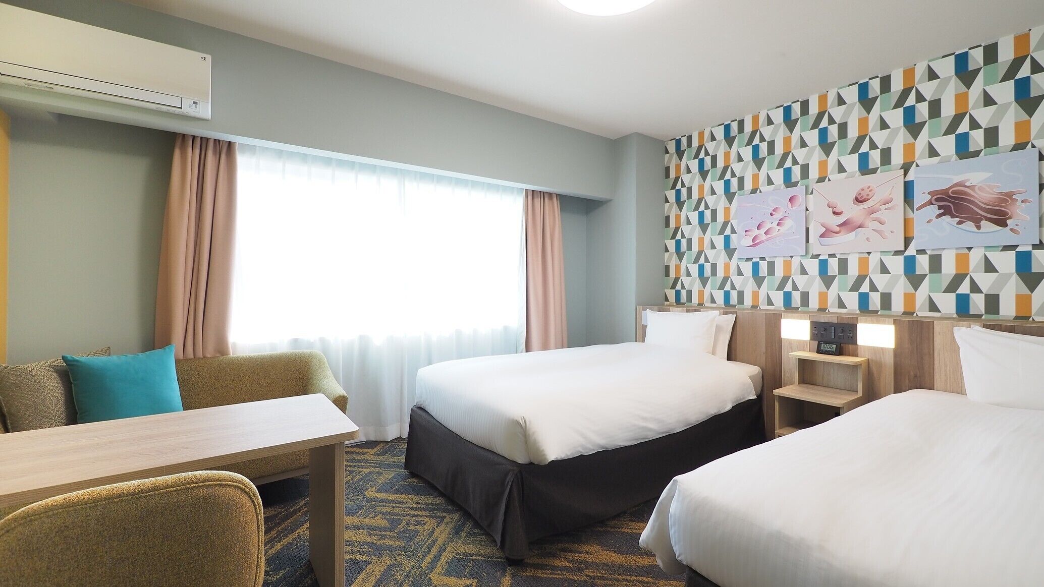 A hotel room with two beds, a desk, a chair, a nightstand, and a wall with geometric patterned wallpaper.