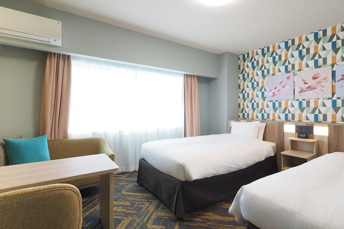 Une chambre d’hôtel avec deux lits, un bureau, une chaise, une table de chevet et un mur recouvert d’un papier peint à motifs géométriques.