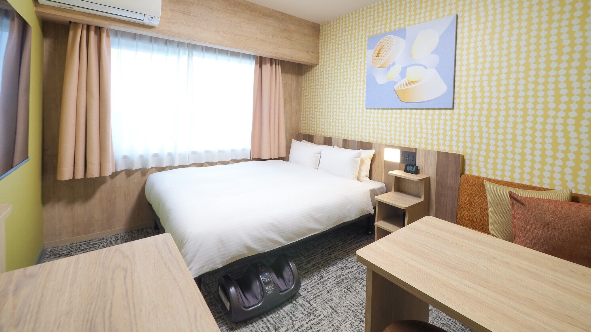 A hotel room with a bed, bedside table, chair, and a wall-mounted air conditioner.