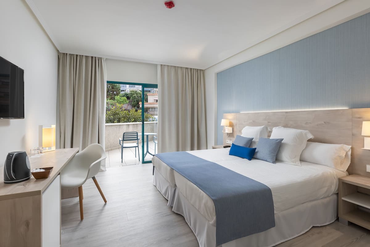A modern hotel room with a large bed, a desk with a coffee maker, a chair, and a view of the outdoors.