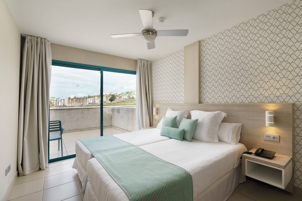 A hotel room with a bed, a nightstand with a phone, a ceiling fan, and a view of the outdoors.