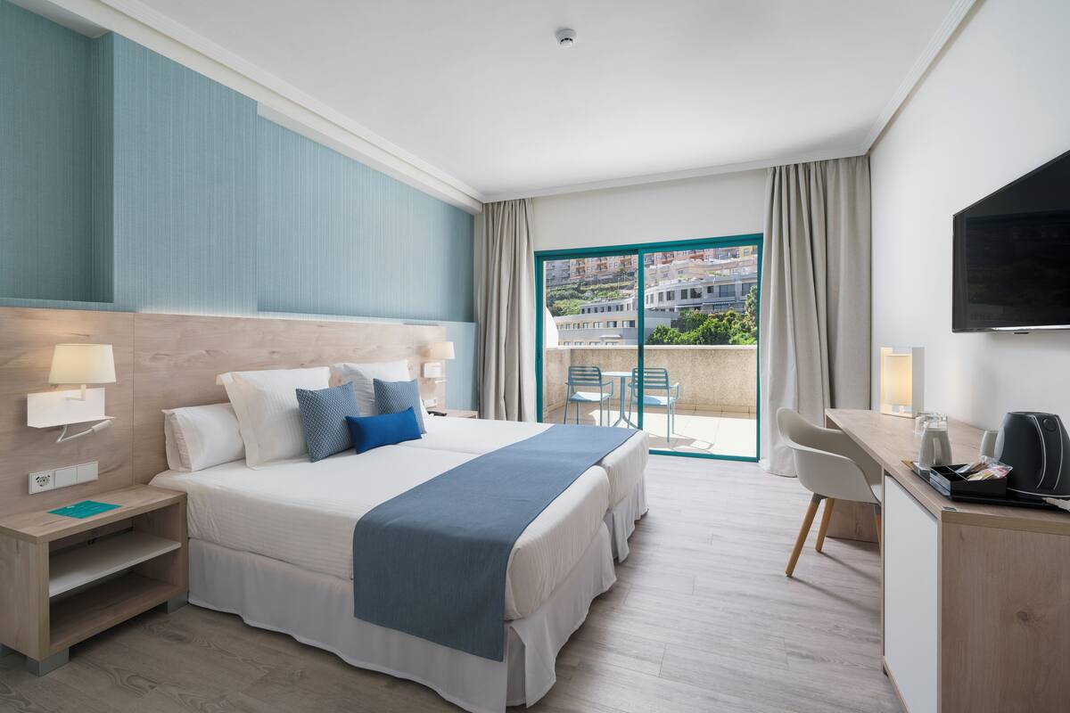 A hotel room with a bed, desk, and a view of buildings through a large window.